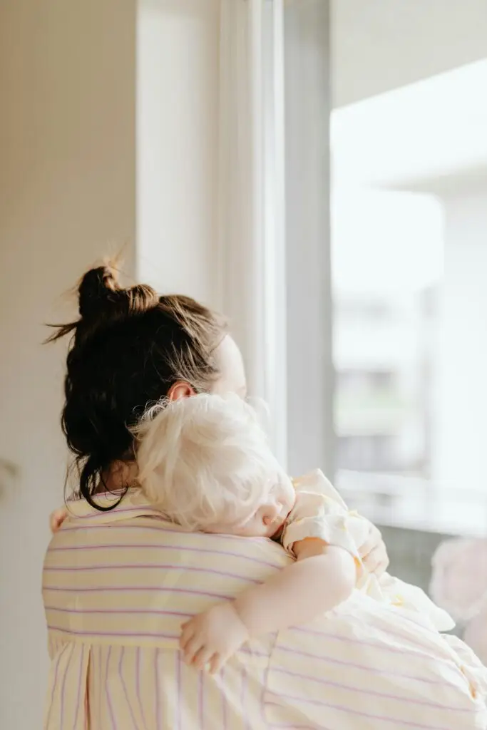 A mother gently holds her sleeping child near a sunlit window.